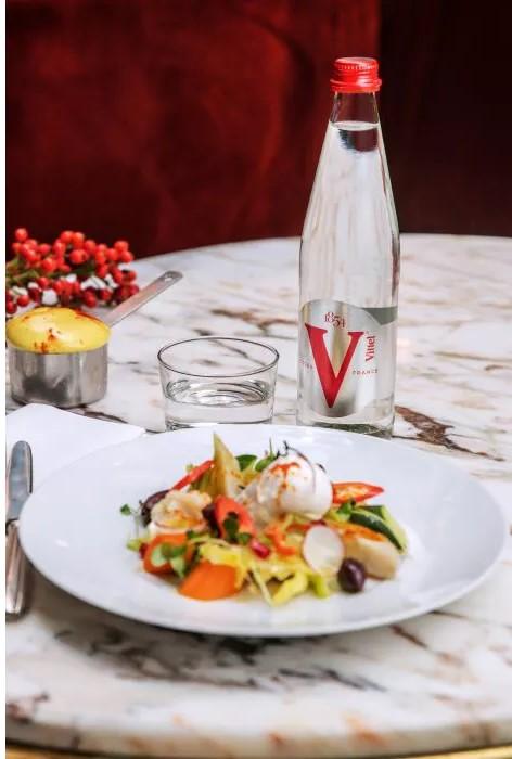 Une salade colorée avec un œuf poché, des carottes, des radis et des légumes verts sur une assiette blanche. À côté, une bouteille d'eau Vittel.